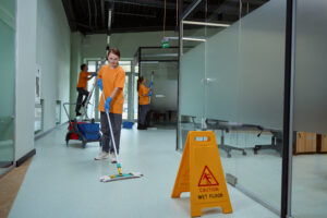 Young Woman Wiping Floor With Wet Mop Near Warning Sign